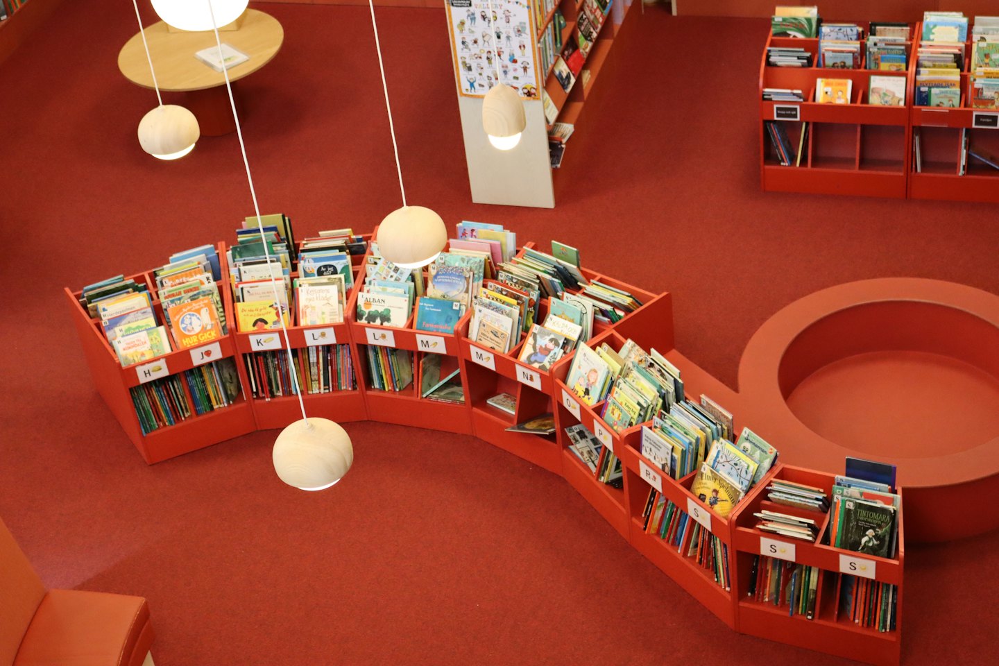 Zakrzywione czerwone regały w dziecięcej bibliotece z okrągłymi miejscami do siedzenia.